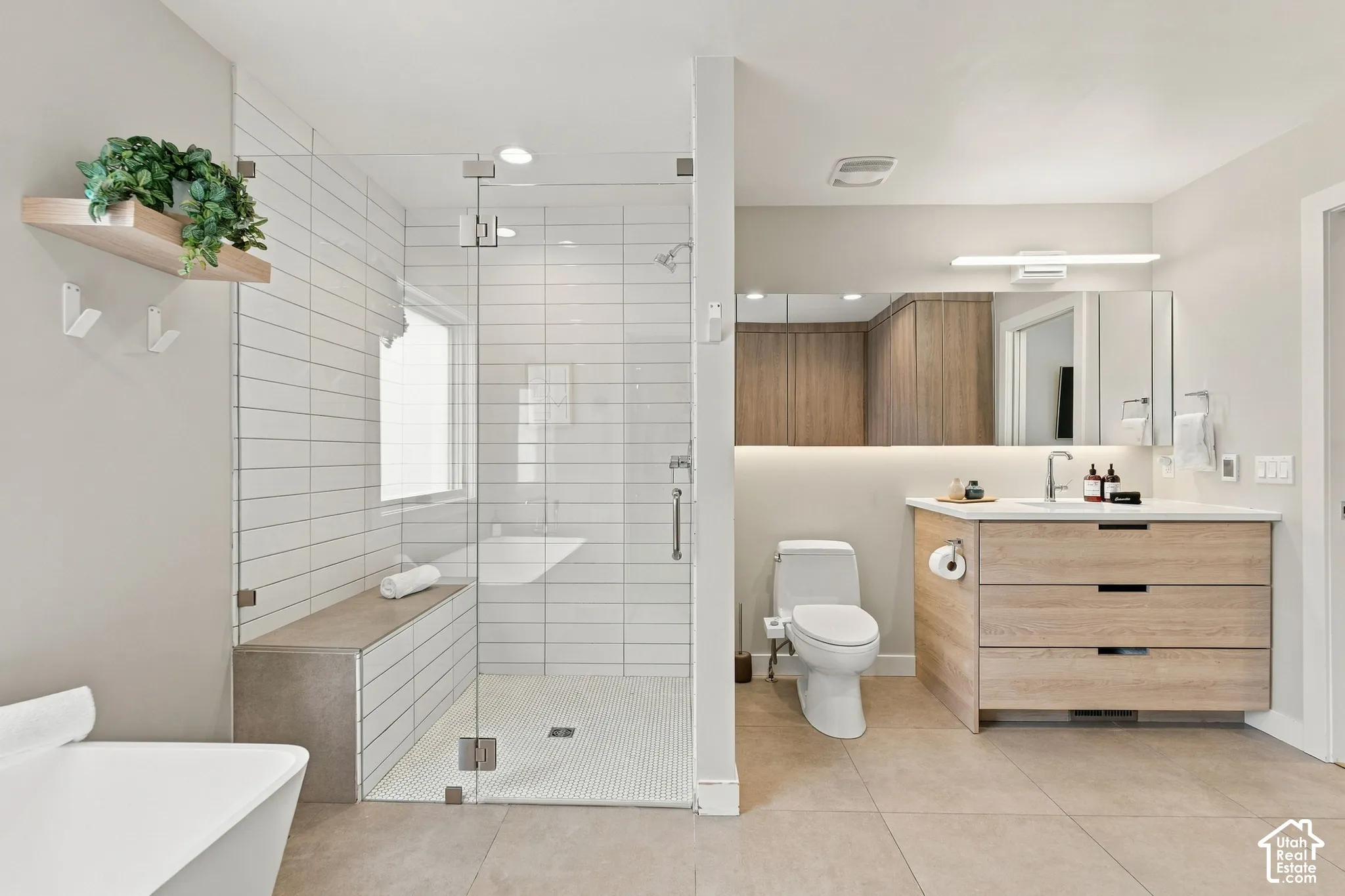 Bathroom featuring a glass-enclosed shower with horizontal wall tile and a built-in bench, a wood-finish vanity with a stone countertop, a full-width mirror with integrated lighting, and neutral floor tile