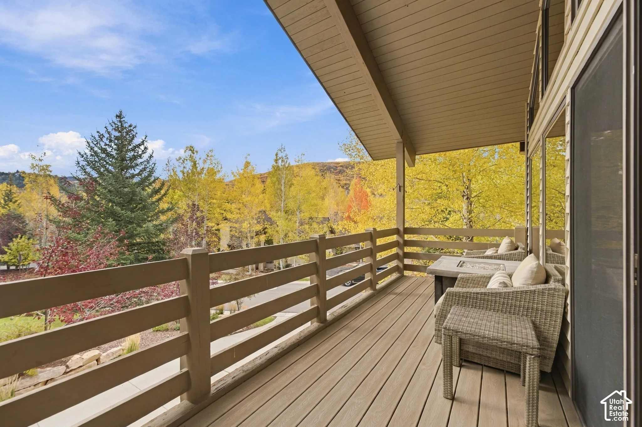 Covered deck featuring horizontal wood-finish railings, composite plank decking, and a sloped beadboard ceiling, overlooking mature deciduous and coniferous trees