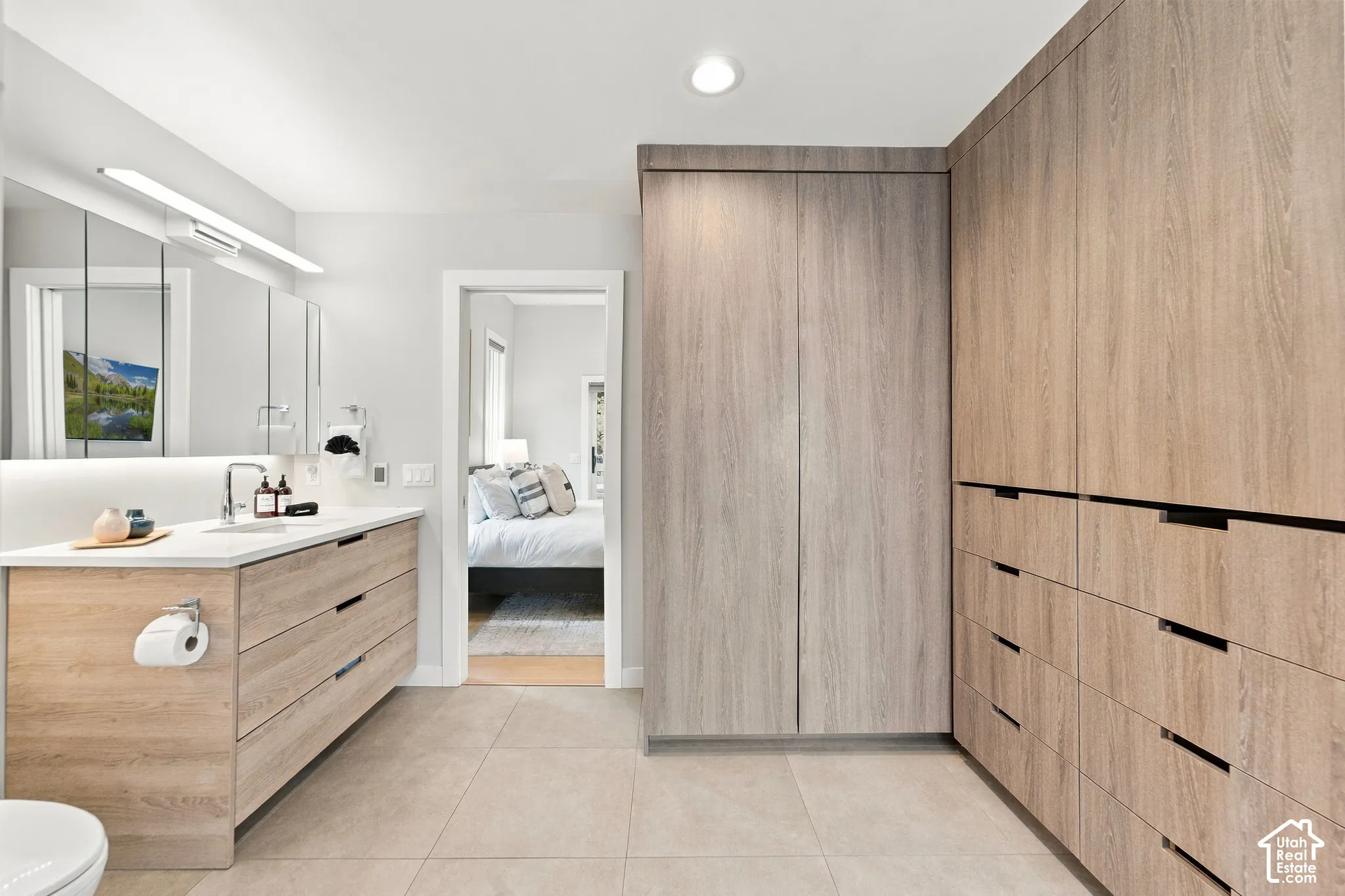 Wood-finish vanity with a light-toned countertop, floor-to-ceiling wood-finish built-in cabinetry, large-format tile flooring, recessed ceiling lighting, and a full-width vanity mirror