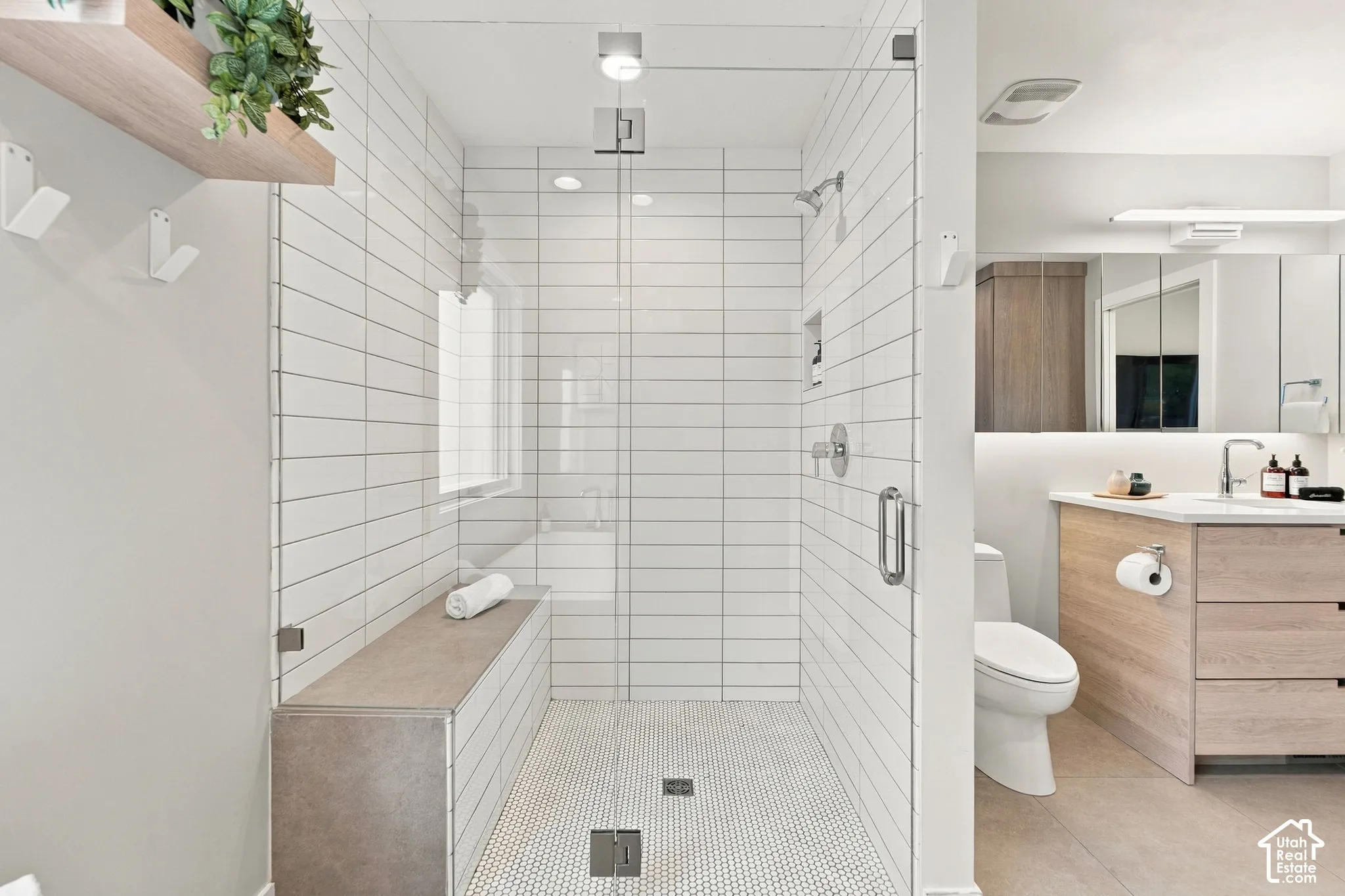 Walk-in shower featuring white subway wall tile, mosaic tile flooring, a built-in tiled bench, and a frameless glass enclosure, alongside a wood-finish vanity with a clean-lined sink and mirror