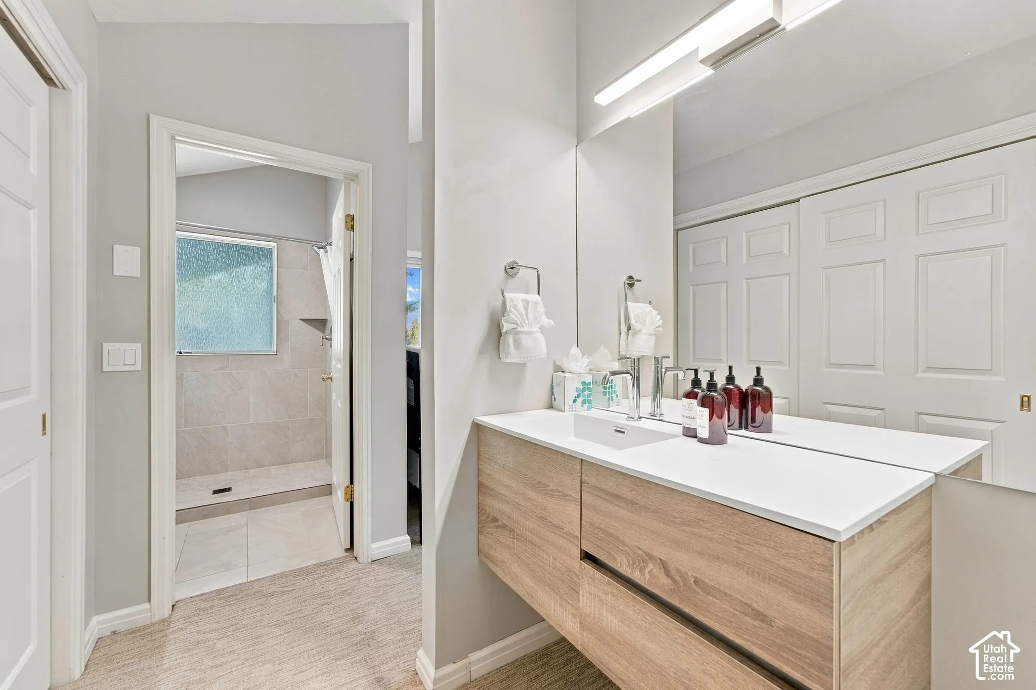 Floating wood-finish vanity with a white countertop and integrated sink, full-width mirror, overhead bar lighting, and tiled walk-in shower