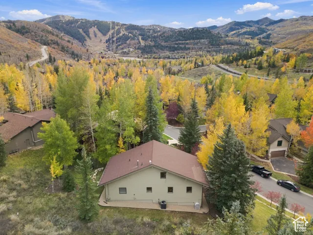Single-story residence featuring a shingled roof, neutral-toned siding, and a concrete patio, set against a backdrop of mature deciduous and evergreen trees with expansive mountain terrain