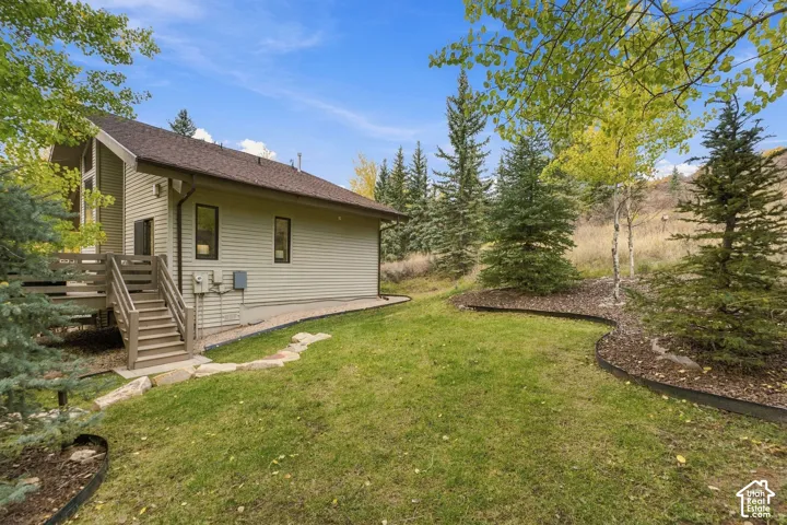 Exterior side elevation featuring horizontal siding, a gabled roof, an attached wooden deck with stairs, stone border edging, and mature coniferous trees