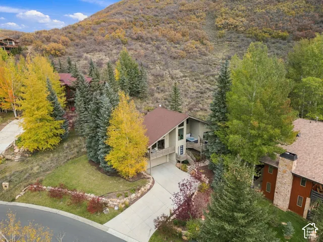 Multi-level residence featuring a pitched roofline, integrated balcony, concrete driveway, stone retaining wall, and mature landscaping set against a mountainous backdrop