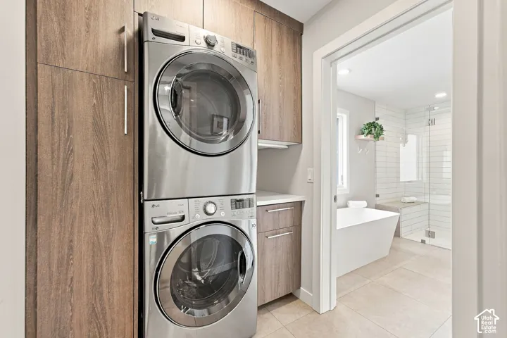 Stacked stainless steel laundry appliances, custom wood-finish cabinetry, stone-surface countertop, and ceramic tile flooring