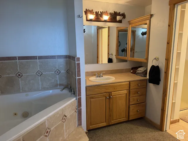 Jetted soaking tub with stone tile surround and decorative inlays, wooden vanity cabinet, integrated sink, and dual-mirror assembly with overhead decorative lighting fixture