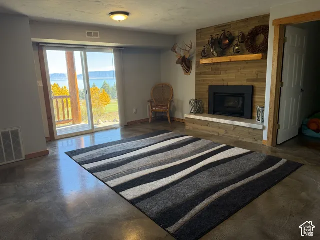 Fireplace featuring a stone-tile hearth, wooden plank surround, and natural wood mantel, complemented by polished concrete flooring and a sliding glass door opening to a wooden deck with mountain and water scenery
