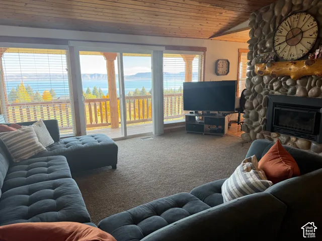 Floor-to-ceiling windows, river-stone fireplace with a natural wood mantel, wood-plank ceiling, and sliding glass doors opening to an exterior deck