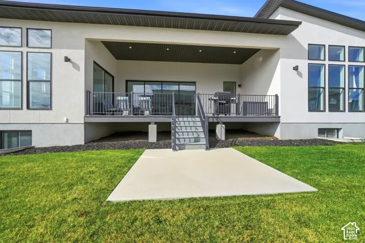 Covered rear deck featuring dark metal railings, stairs to ground level, recessed ceiling lighting, and a dedicated concrete patio slab set within a manicured lawn