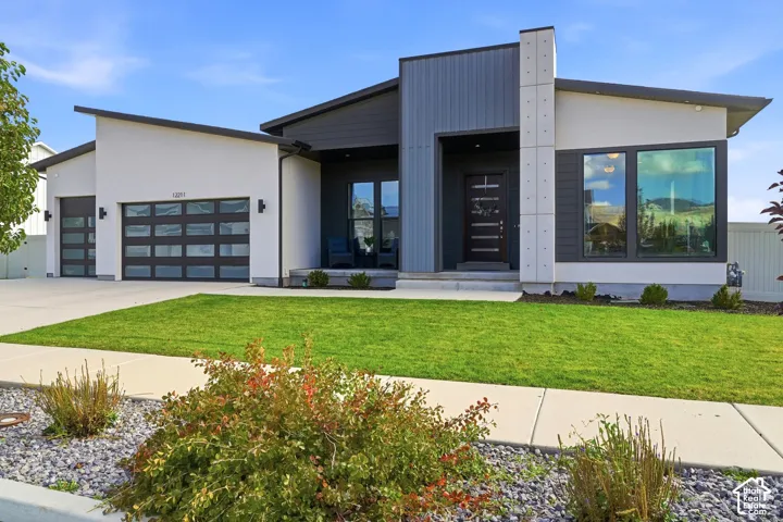 Modern architectural exterior featuring smooth stucco siding, vertical metal paneling, large-format picture windows, an attached multi-car garage with glass-paneled doors, and a decorative concrete chimney stack