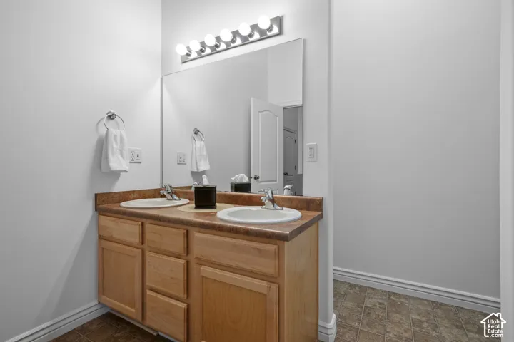 Dual-sink vanity with light-tone wood cabinetry, stone-finish countertop, full-width mirror, and multi-bulb wall lighting