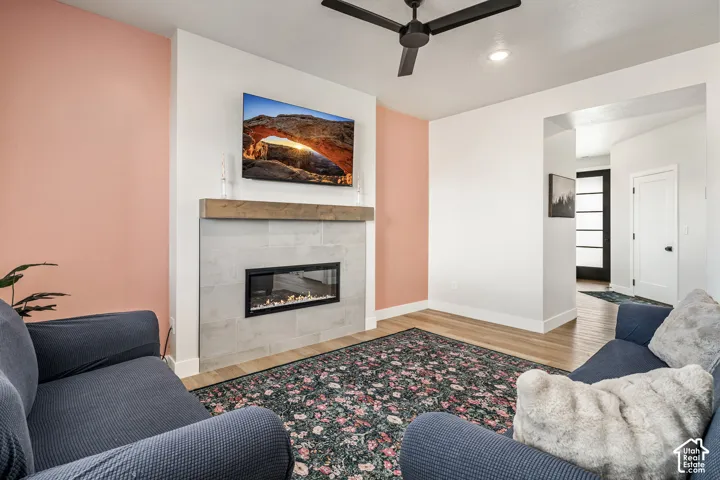 Linear fireplace with a light-toned stone surround, rustic wood mantel, matte black ceiling fan, and wood-finish flooring