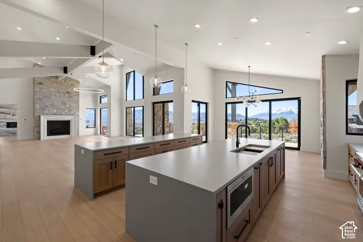 Open-concept kitchen and living area featuring vaulted ceilings with exposed beams, wood-finish flooring, stone fireplace surround, dual kitchen islands with solid-surface countertops, and expansive windows with mountain scenery