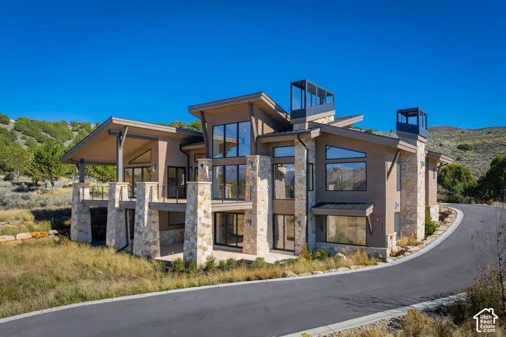 Modern exterior featuring multi-level stone pillars, horizontal wood-finish siding, expansive floor-to-ceiling windows, covered deck space, and distinct architectural roof chimneys
