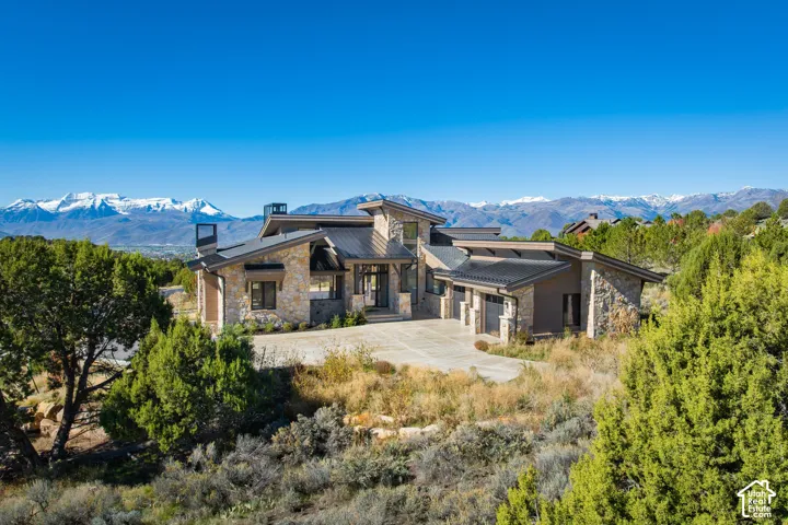 Contemporary architectural exterior featuring natural stone siding, metal roofing, expansive glass windows, and a concrete driveway, set against a backdrop of snow-capped mountain peaks