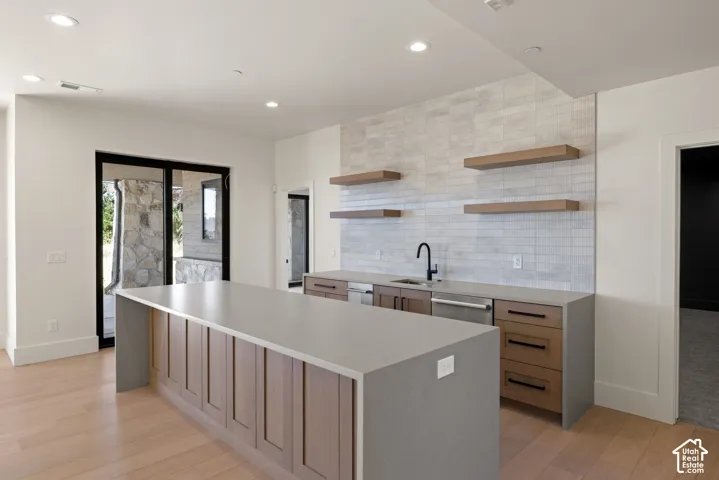 Kitchen featuring a waterfall-edge island with stone-finish countertops, wood-finish cabinetry, floating wood shelving, full-height tile backsplash, and matte black plumbing fixtures