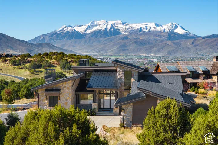 Modern mountain residence featuring stone veneer masonry, dark metal roof accents, multi-level rooflines, floor-to-ceiling glass windows, and a prominent backdrop of snow-capped mountains