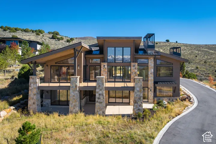 Modern residence featuring multi-level stone pillars, floor-to-ceiling windows, expansive elevated decking with metal railing, and a multi-pitch roofline