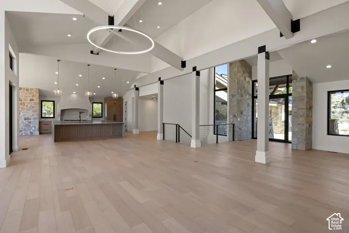 Vaulted ceilings with exposed structural beams, wide-plank wood-finish flooring, stone accent pillars, custom kitchen cabinetry with large island, and contemporary geometric chandelier
