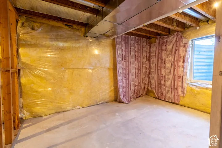 Unfinished basement space featuring exposed wood framing, batt insulation with a vapor barrier, metal ductwork, and an egress-style window with a corrugated metal well