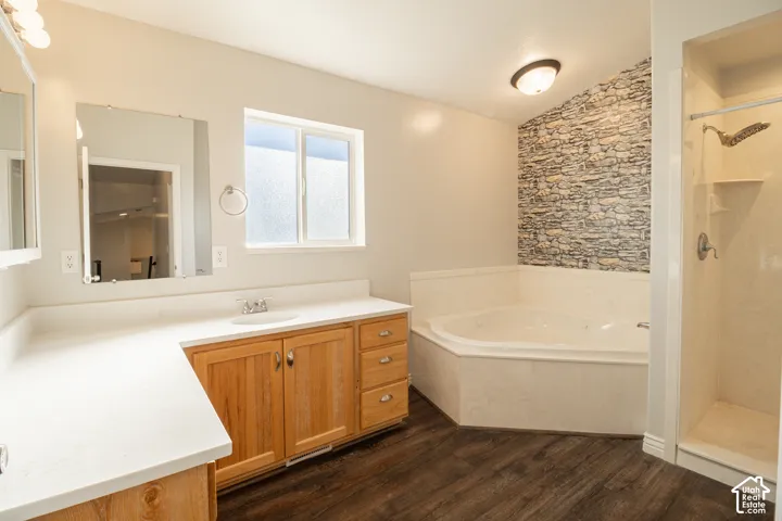 Bathroom featuring wood-finish flooring, a jetted soaking tub with a stone-textured accent wall, a separate shower stall, and a vanity with a white countertop