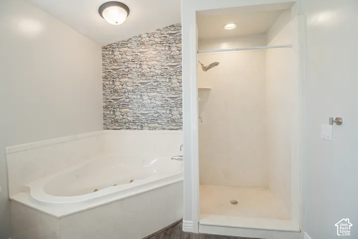 Jetted corner tub with stone-textured accent wall, separate stall shower with recessed lighting, and wood-finish flooring