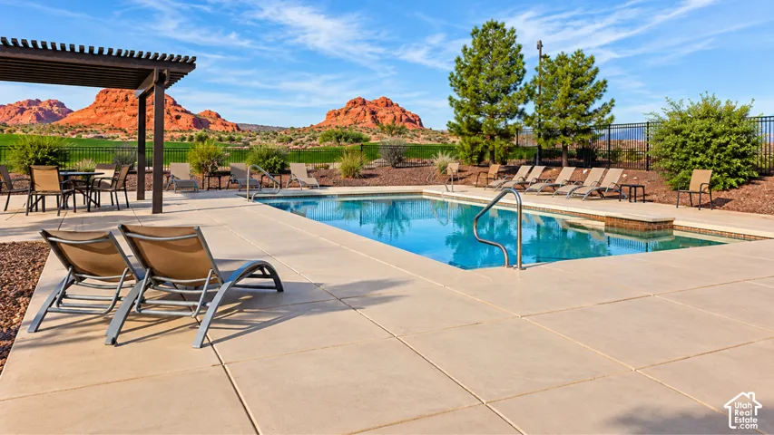 Rectangular inground pool with a stone-accented edge, expansive concrete patio, overhead wooden pergola, metal perimeter fencing, and scenic red rock formations