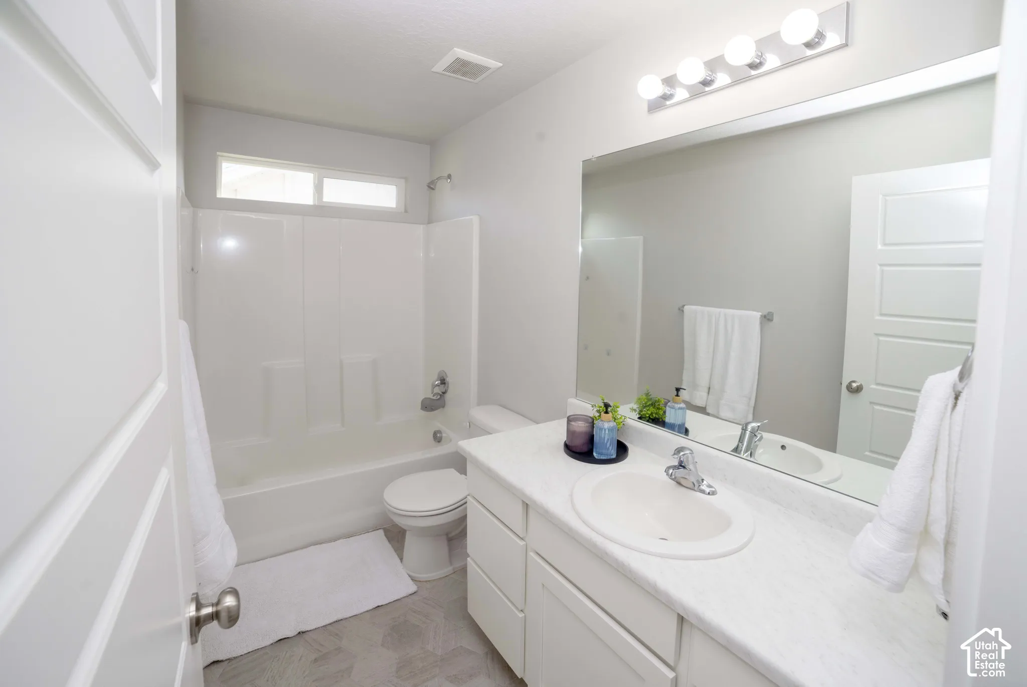 Full-width vanity with solid-surface countertop and integrated sink, large wall-mounted mirror, four-bulb light fixture, tub-shower combination with surround, and patterned flooring