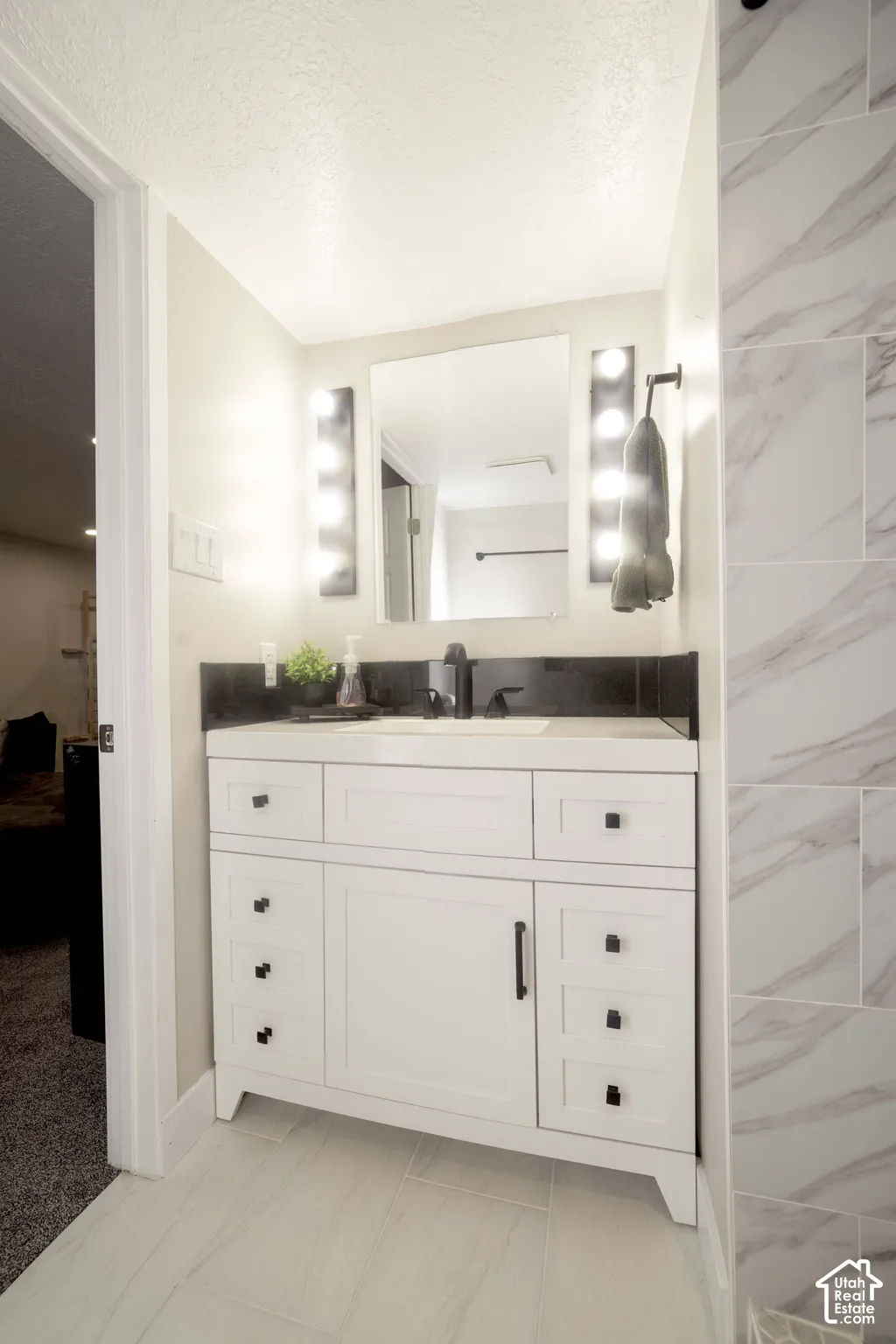 White vanity with dark hardware, stone-finish backsplash, rectangular undermount sink, wall-mounted vanity lighting, and light-toned tile flooring