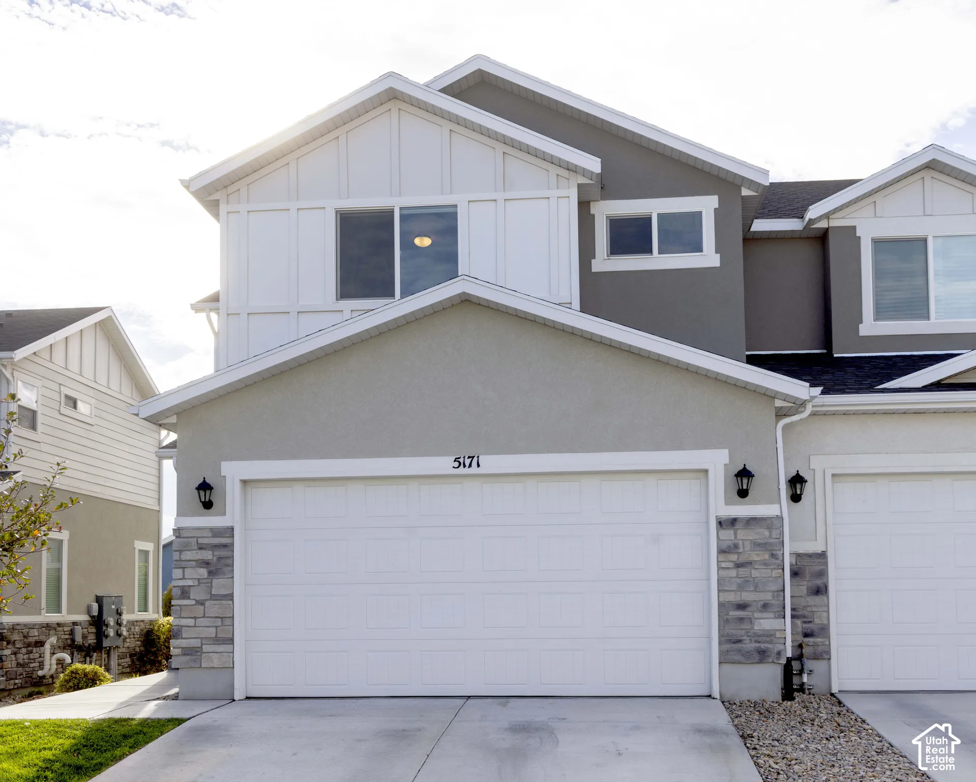 Exterior featuring board-and-batten siding, neutral stucco finishes, stone veneer accents, an attached two-car garage, and wall-mounted exterior light fixtures