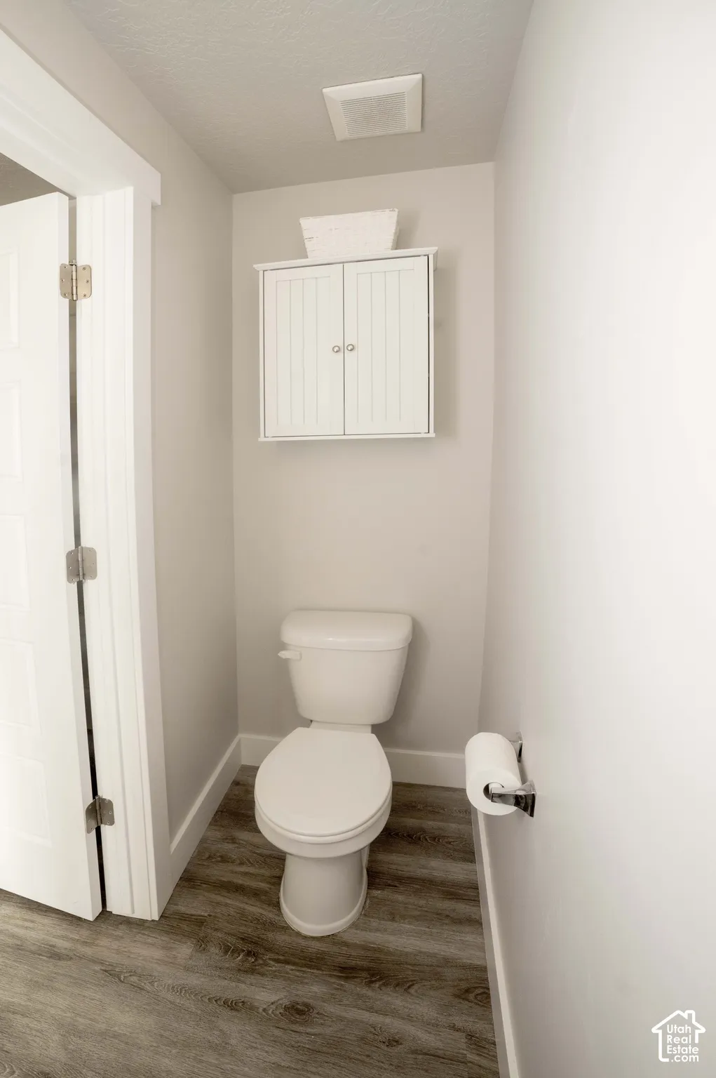 Neutral-toned space featuring LVP flooring, a white porcelain toilet, wall-mounted white shaker-style cabinetry, and polished chrome hardware