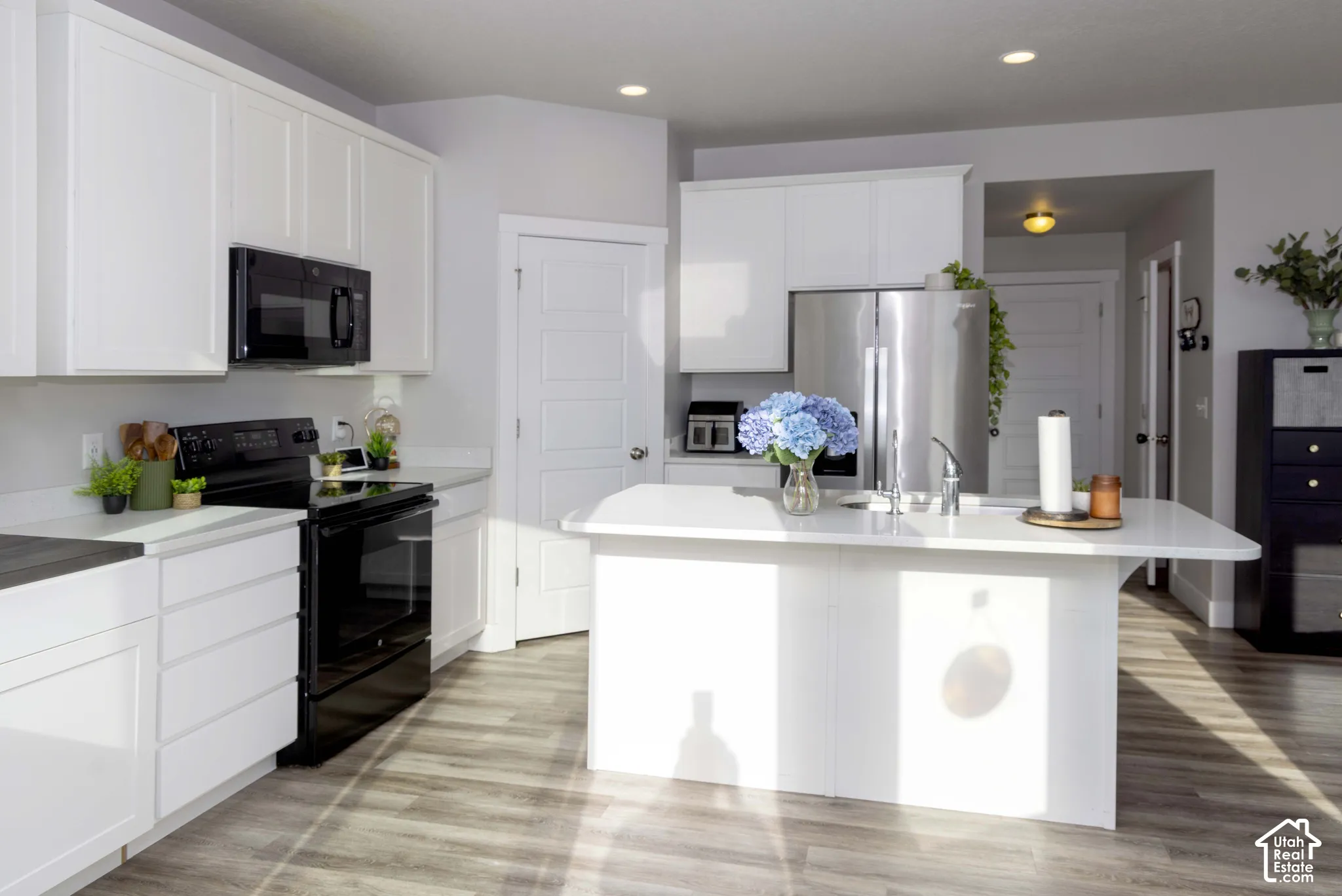 Kitchen featuring white shaker-style cabinetry, quartz countertops, a central island with an integrated sink, stainless steel appliances, and LVP flooring