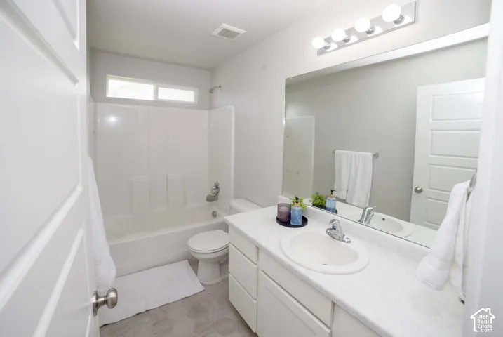 Full-width vanity with solid-surface countertop and integrated sink, large wall-mounted mirror, four-bulb light fixture, tub-shower combination with surround, and patterned flooring