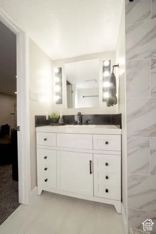 White vanity with dark hardware, stone-finish backsplash, rectangular undermount sink, wall-mounted vanity lighting, and light-toned tile flooring