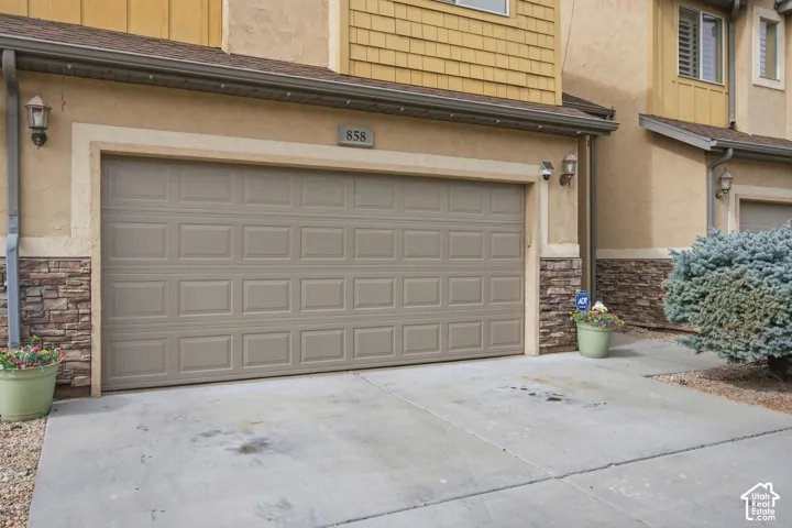 Multi-level exterior featuring a two-car garage with a paneled door, tan stucco siding, decorative stone veneer wainscoting, and shingle-style upper siding accents