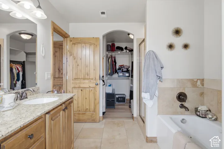 Bathroom featuring a stone-finish countertop with an undermount sink, natural wood-finish cabinetry, arched-top wood doors, tile flooring, and a tiled soaking tub surround with bronze-finish fixtures