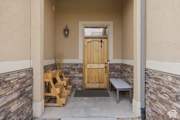 Covered entryway featuring a natural wood-finish front door with a transom window, complemented by stone veneer wainscoting and a wall-mounted light fixture