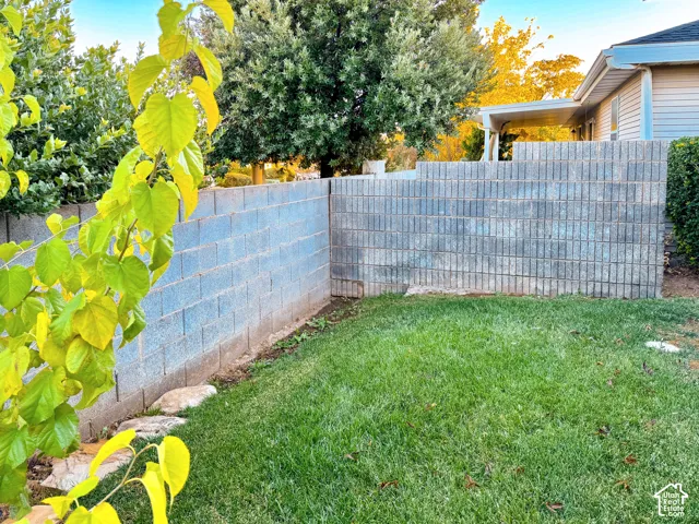 Backyard featuring a tiered concrete block perimeter wall, natural grass lawn, mature trees, and a covered patio structure