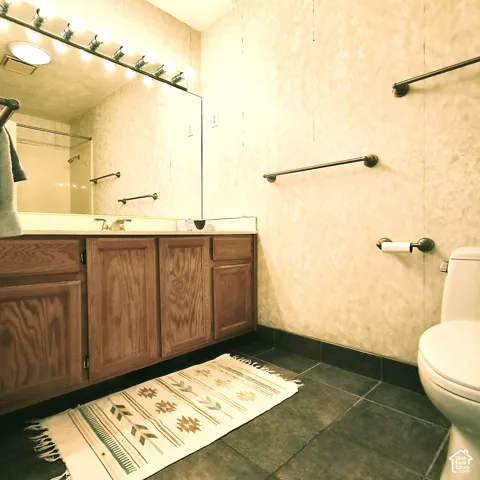 Bathroom featuring wooden cabinetry, a wall-to-wall vanity mirror with integrated multi-bulb lighting, dark-toned tile flooring, and metal wall-mounted hardware