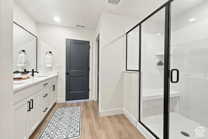Bathroom featuring a dual-sink vanity with white cabinetry, quartz countertop, matte black hardware, glass-enclosed shower with a built-in bench, and wood-finish flooring