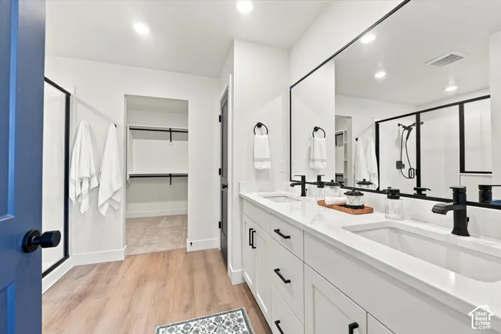Dual-sink vanity with white cabinetry, light-toned countertop, and matte black hardware, accompanied by a glass-enclosed shower, wall-to-wall vanity mirror, and walk-in closet space with built-in shelving