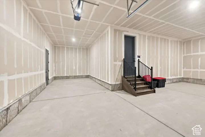 Garage interior featuring concrete flooring, taped drywall, recessed lighting, and a staircase with a black metal railing