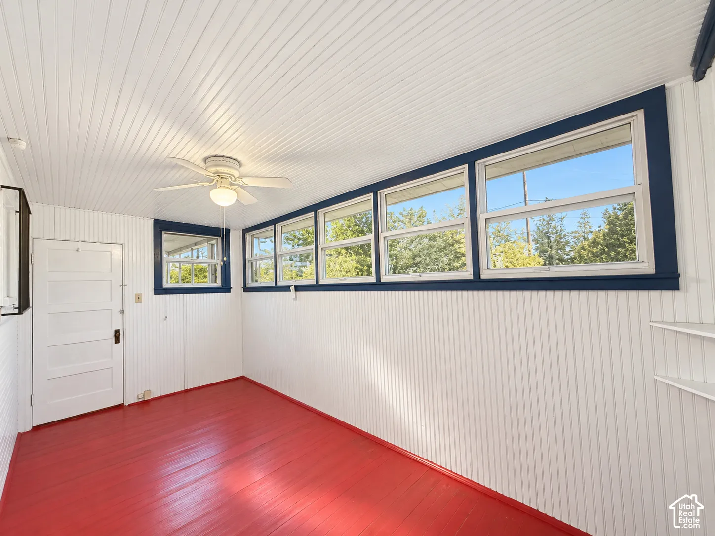 Enclosed porch featuring vertical beadboard paneling, painted wood-finish flooring, a ceiling-mounted fan with light fixture, and multiple windows with contrasting trim