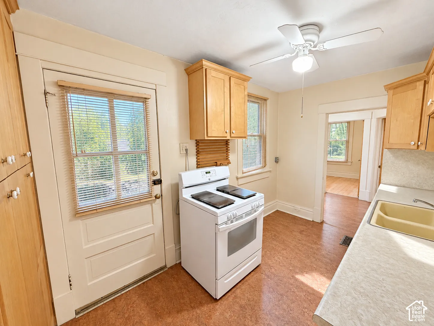 Kitchen featuring light wood-finish cabinetry, a white freestanding range, a dual-basin sink with a solid-surface countertop, and a ceiling-mounted fan with integrated light