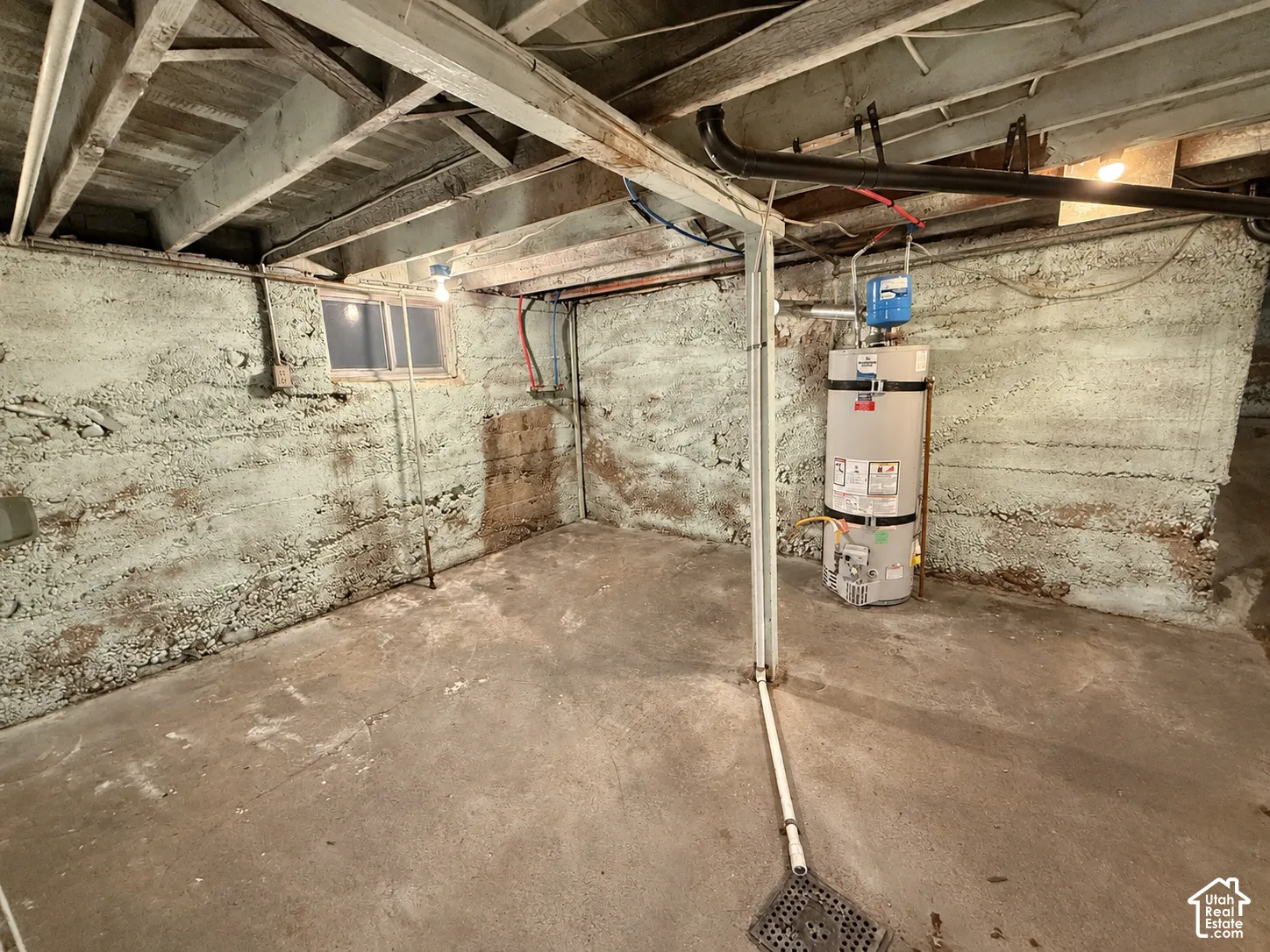 Unfinished concrete basement area featuring exposed ceiling joists, concrete foundation walls, a floor drain, and a vertical water heater