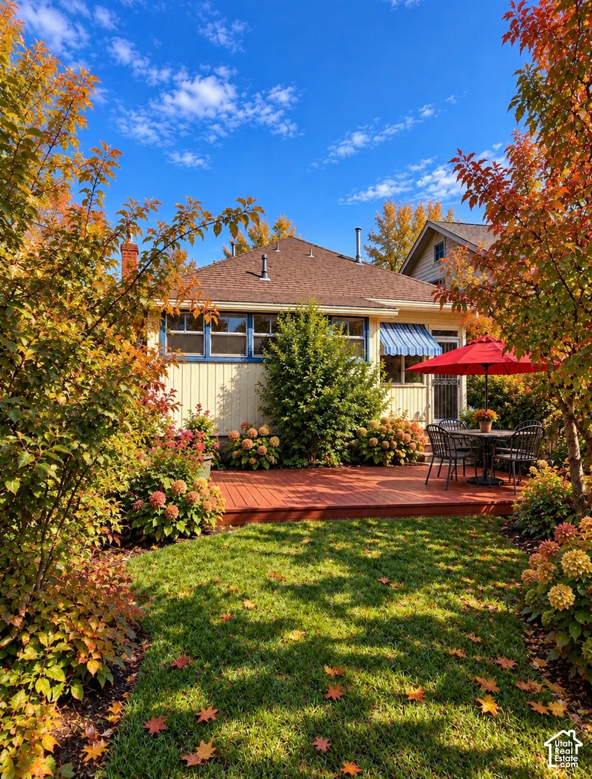 Rear exterior featuring vertical siding, a raised wood deck, a shingled roof, and manicured landscaping with established shrubbery and mature trees