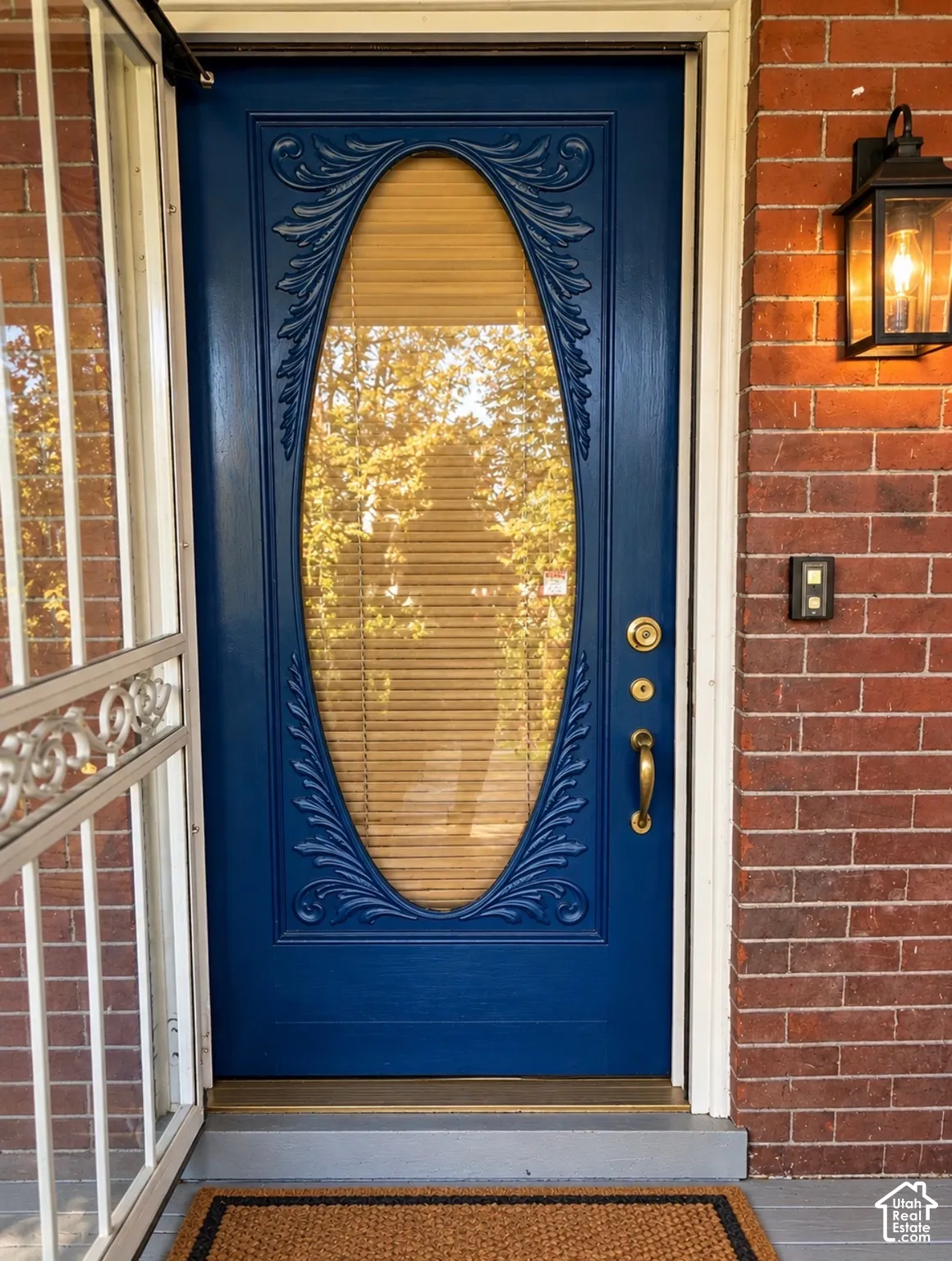 Blue entry door featuring a central oval glass insert with ornate carved detailing, brass-tone hardware, and an exterior lantern-style wall sconce set against a brick facade