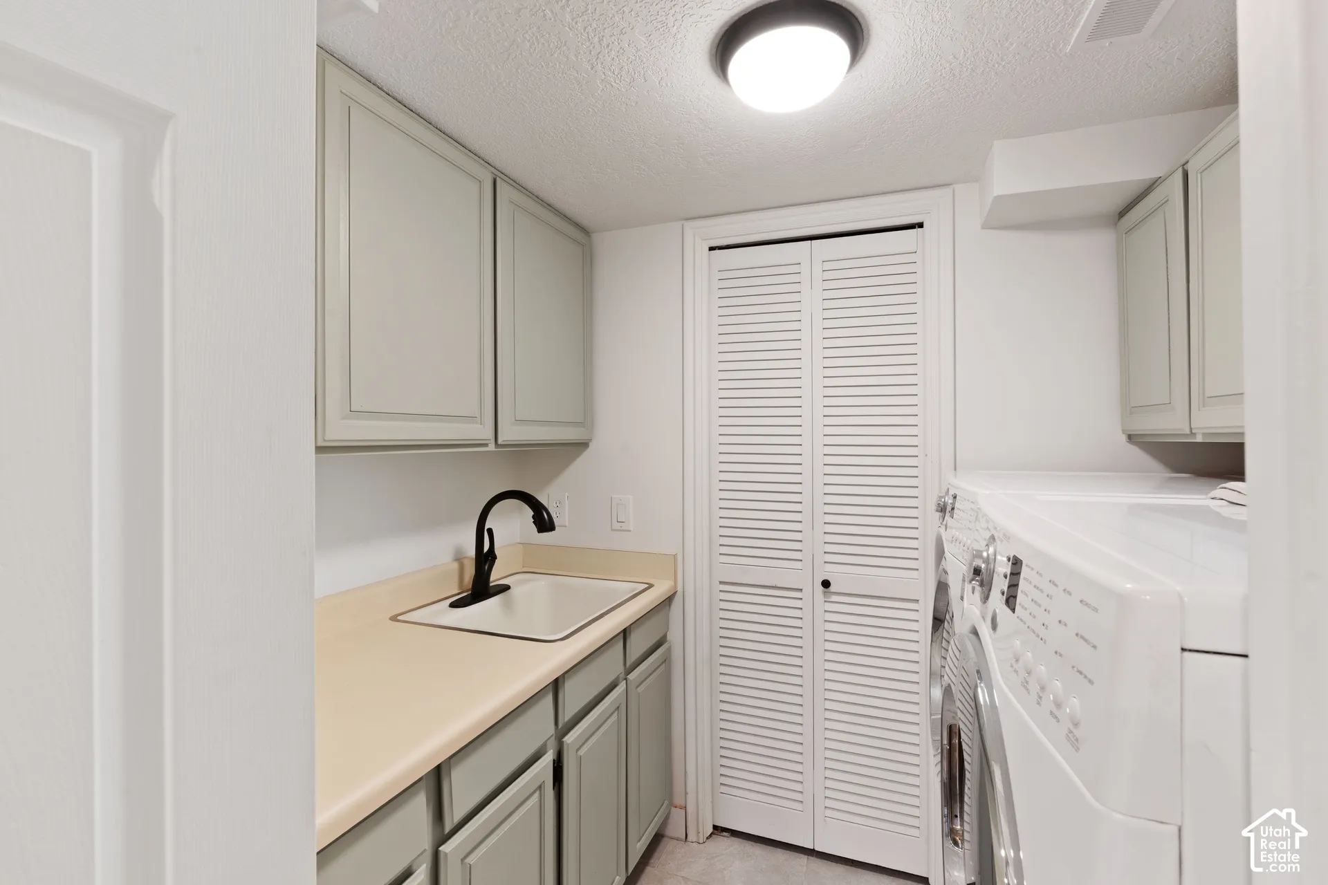 Laundry area featuring a drop-in sink with a matte black faucet, solid surface countertop, light-toned shaker-style cabinetry, and white louvered bifold closet doors