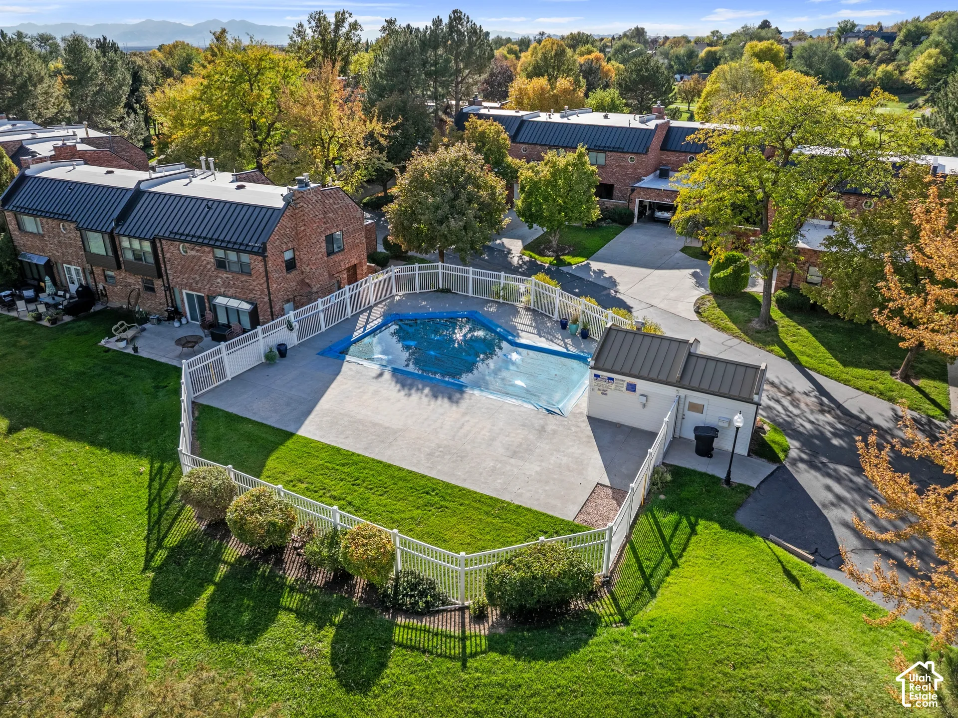 Fenced community swimming pool with surrounding concrete patio and utility structure, set against a backdrop of mature trees and distant mountain range