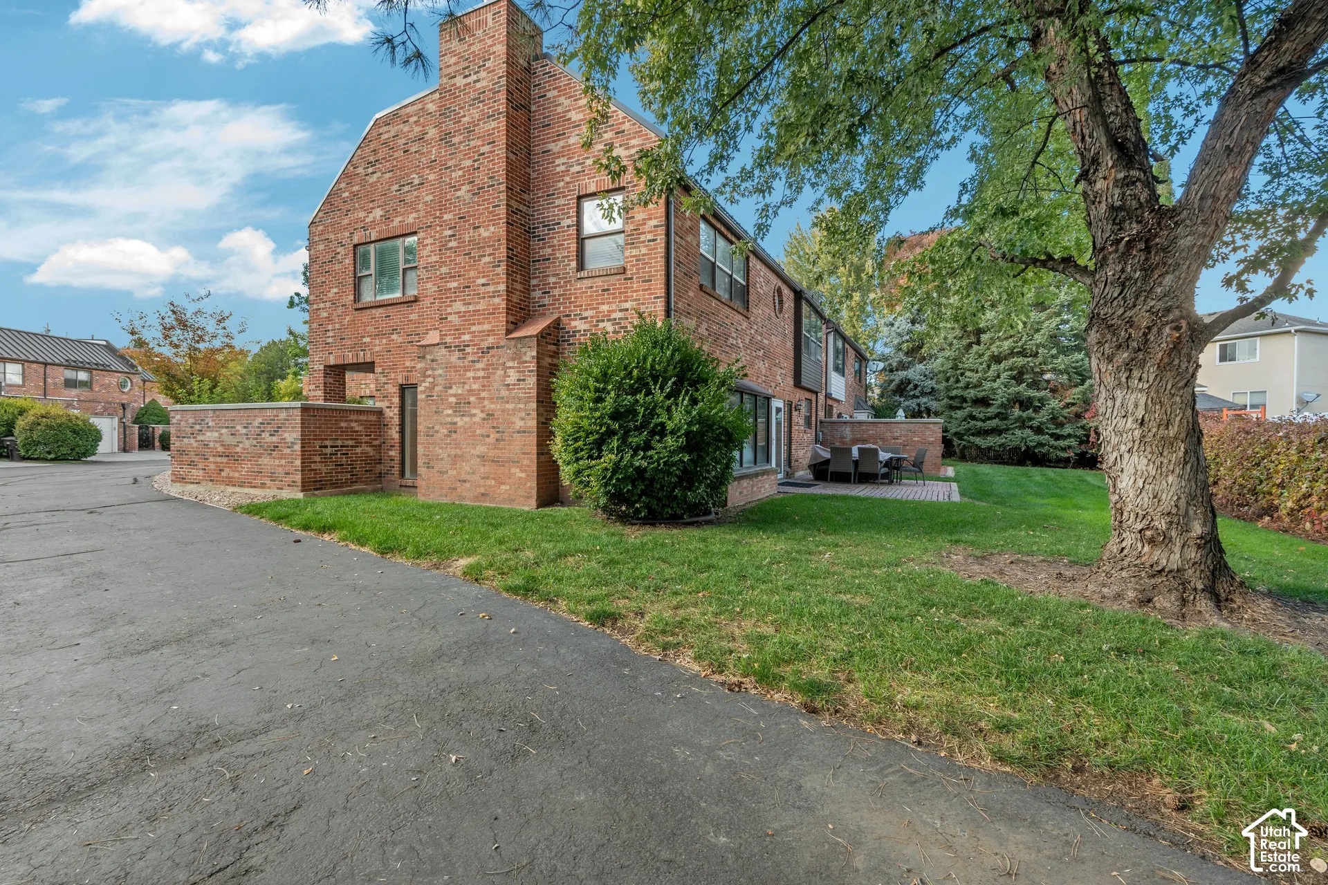 Exterior featuring a multi-story red brick facade, chimney, paved driveway, brick privacy wall, and green landscaping with mature trees